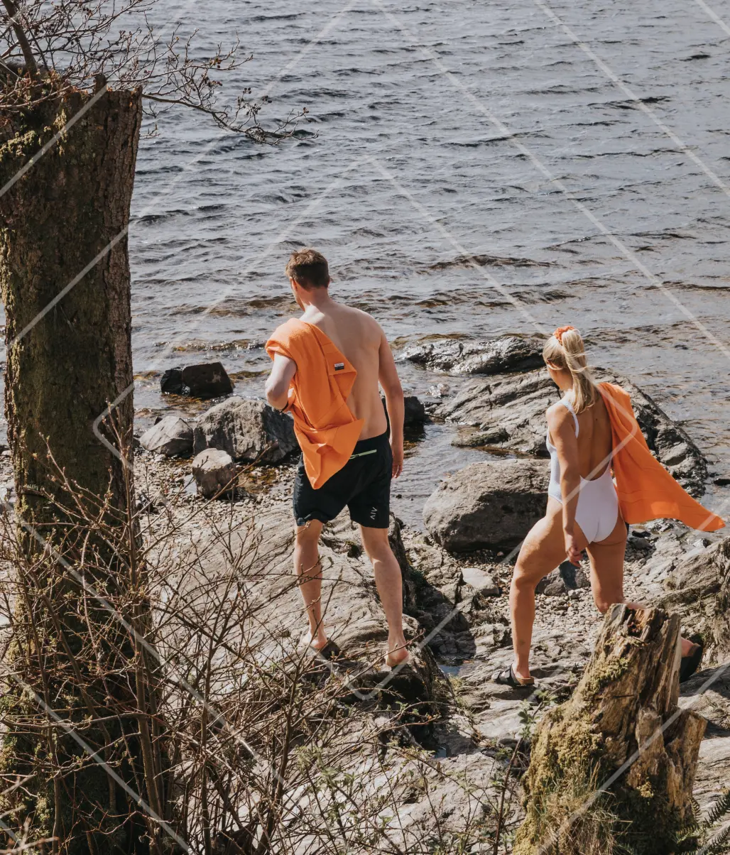 Rona McMillan and her brother walking toward a loch for a wild swim, orange towels over their shoulders, Ben Lomond Gin collab